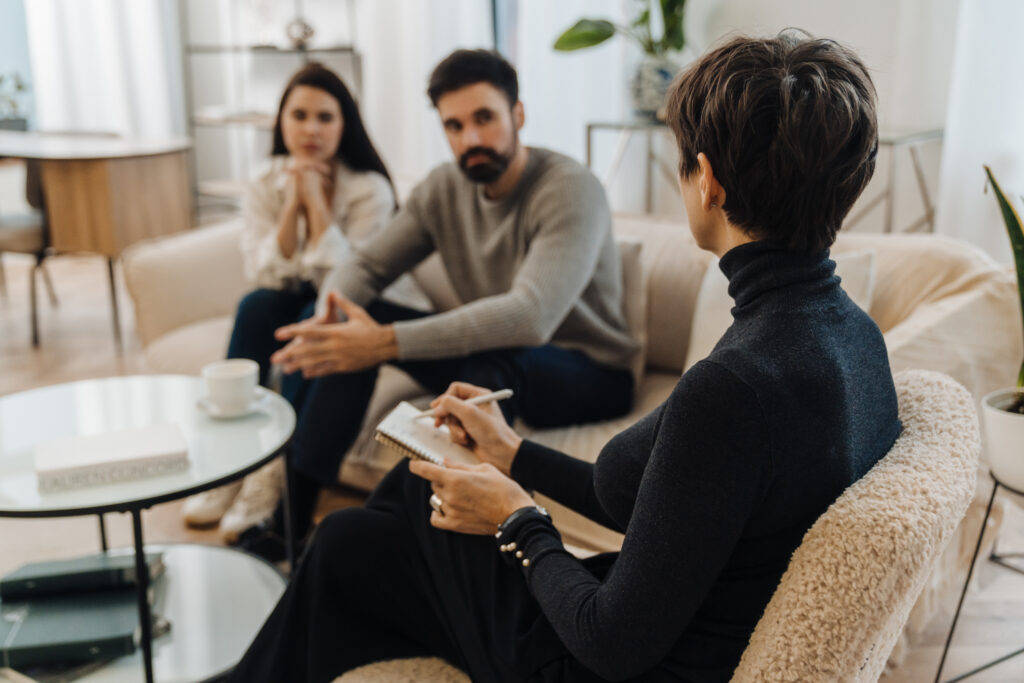 Adult married couple sitting on sofa and having conversation with family psychologist during therapy session at Emberhaven in North Carolina